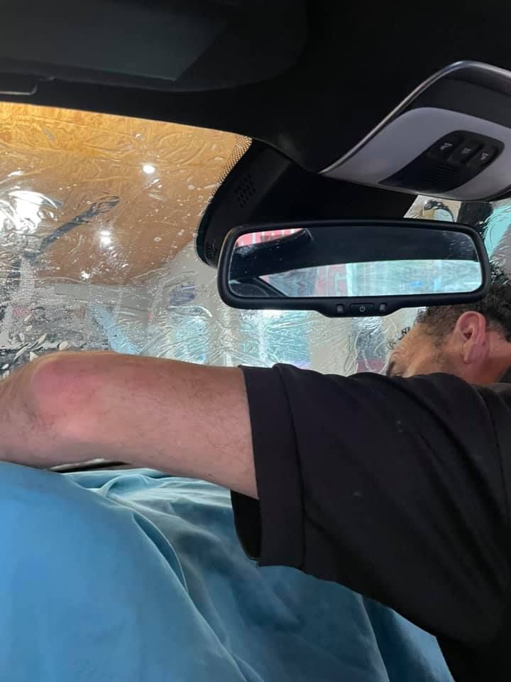 Person working on a car's windshield, inside view. Man in black shirt, using tools. Interior is visible.