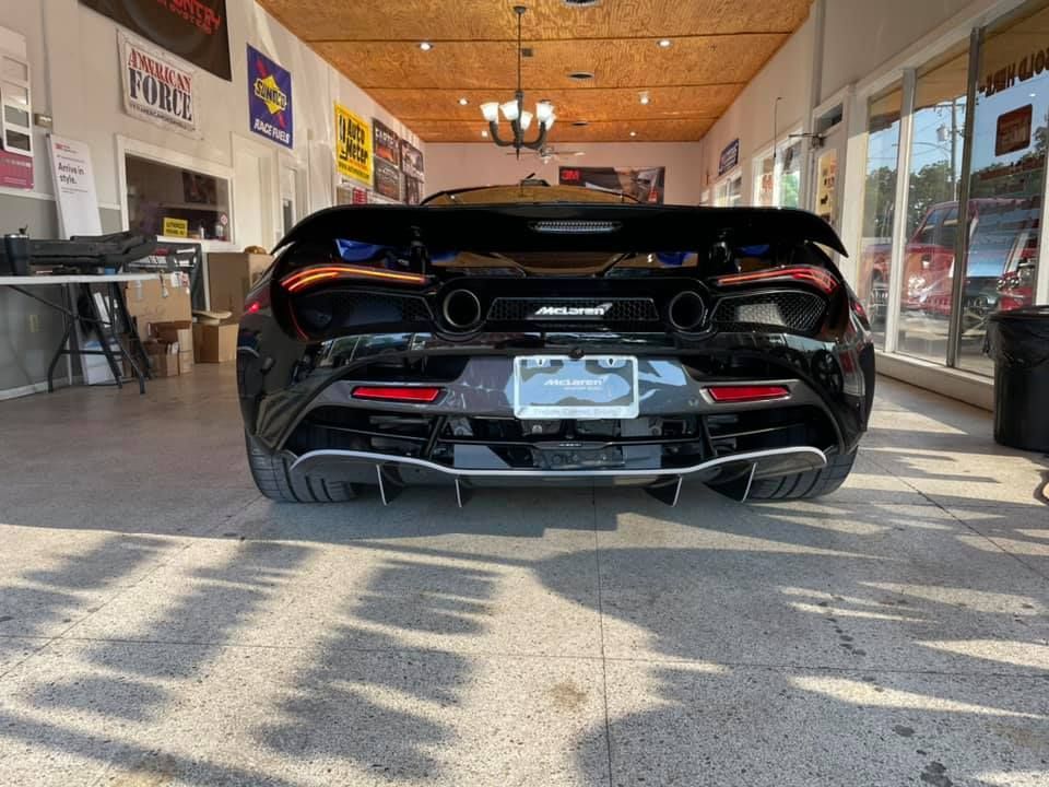 Black McLaren sports car in a shop, rear view.  Silver exhaust tips, license plate, taillights, and large spoiler.