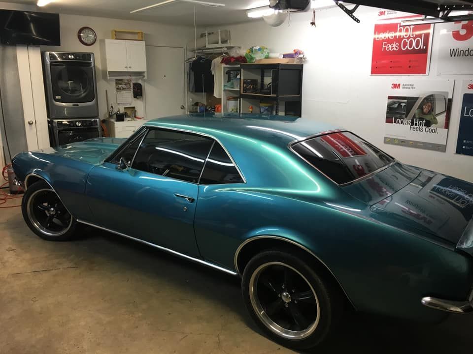 Teal classic Camaro in garage. Black wheels, silver trim. Posters on wall, laundry machines in background.
