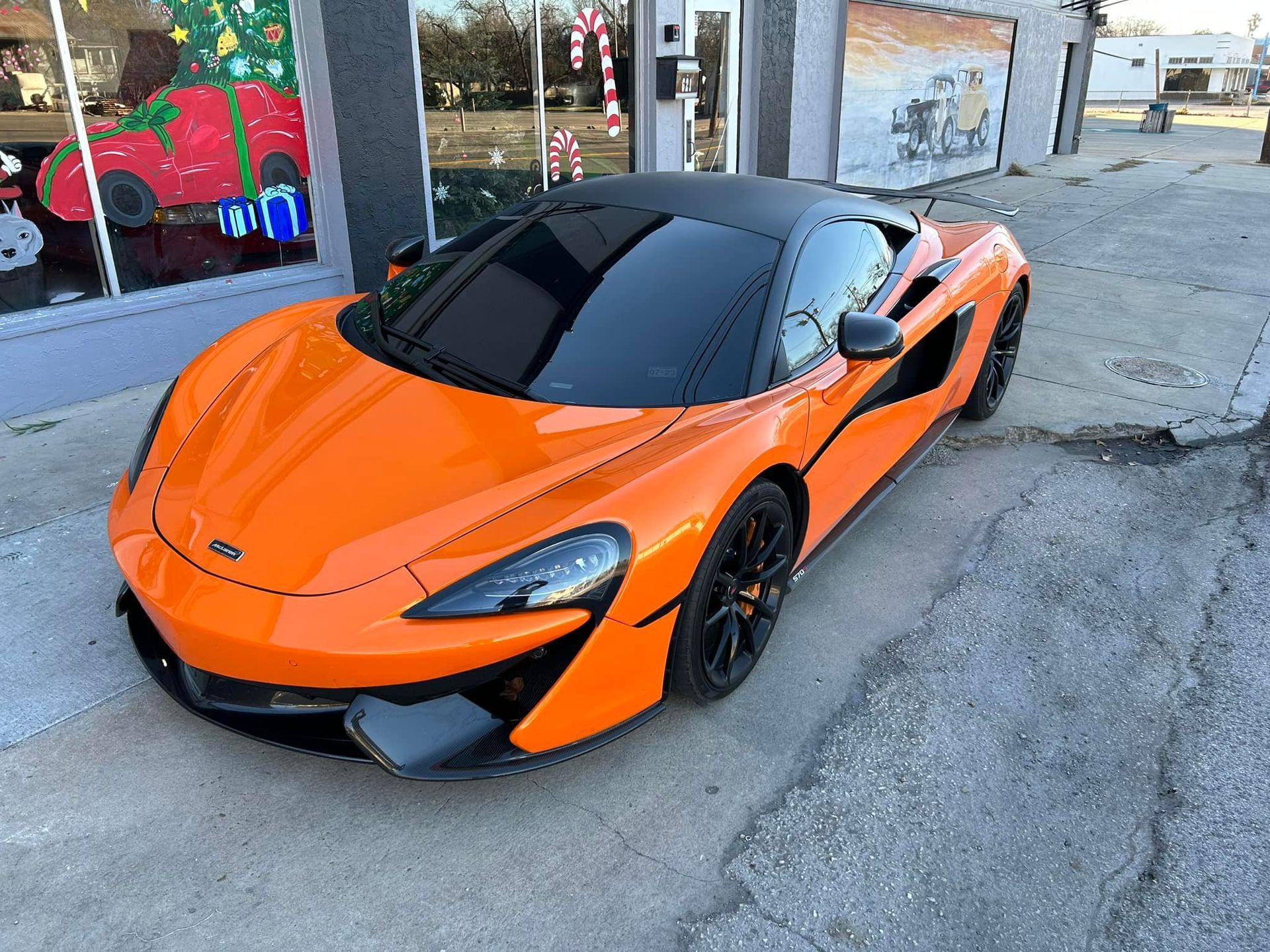 Bright orange McLaren sports car parked on a city street. Black roof, dark wheels, storefront in the background.