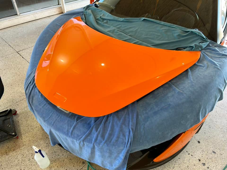 Bright orange car hood resting on a car covered with a blue cloth, in a workshop setting.