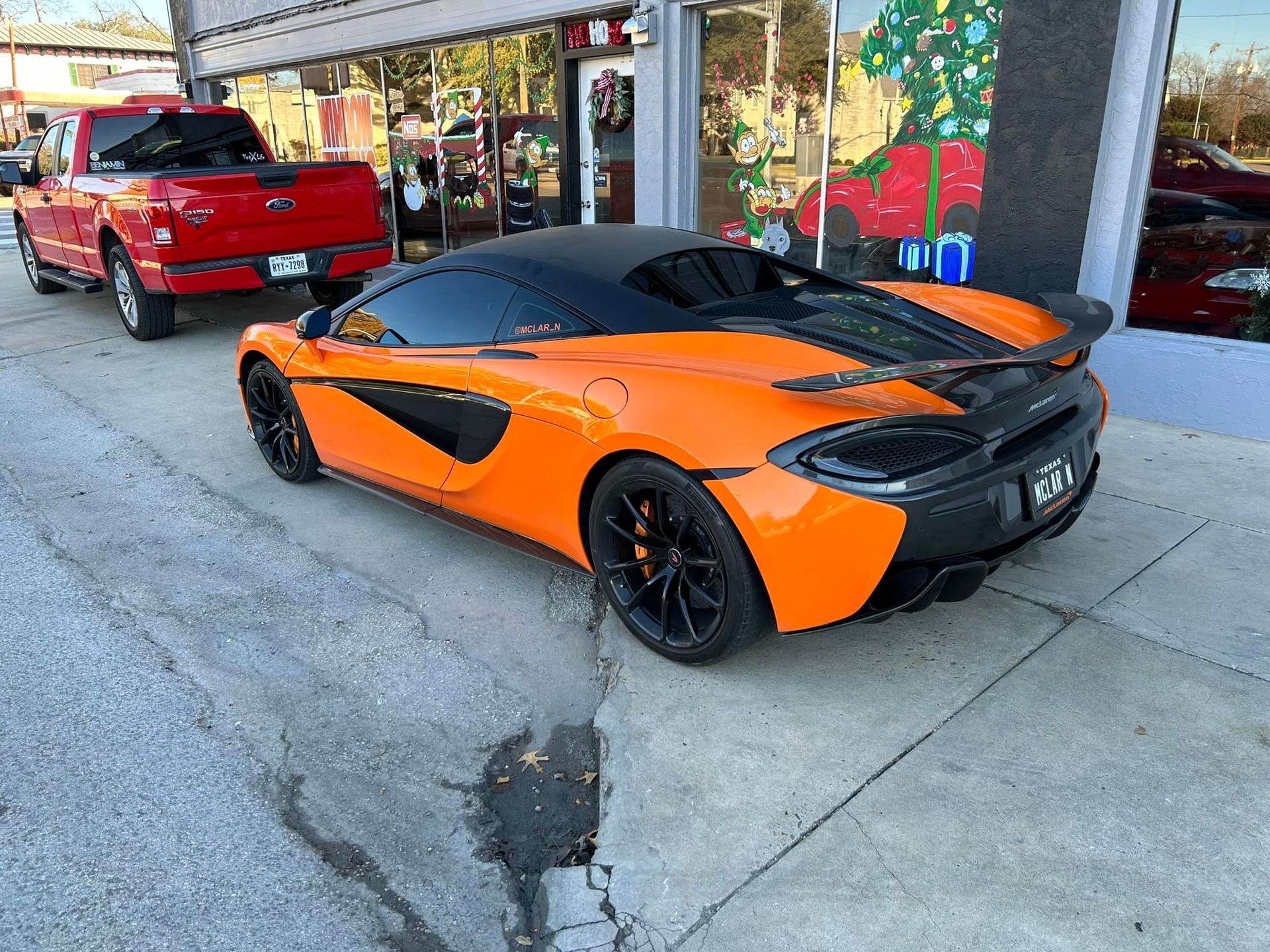 Orange McLaren sports car parked outside a shop, next to a red pickup truck.
