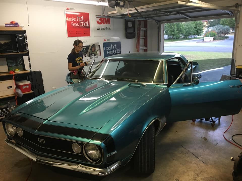A person tinting windows of a teal classic Chevy Camaro in a garage.