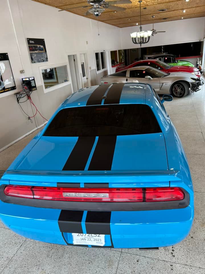 Blue Dodge Challenger with black stripes in a garage, other cars visible in the background.