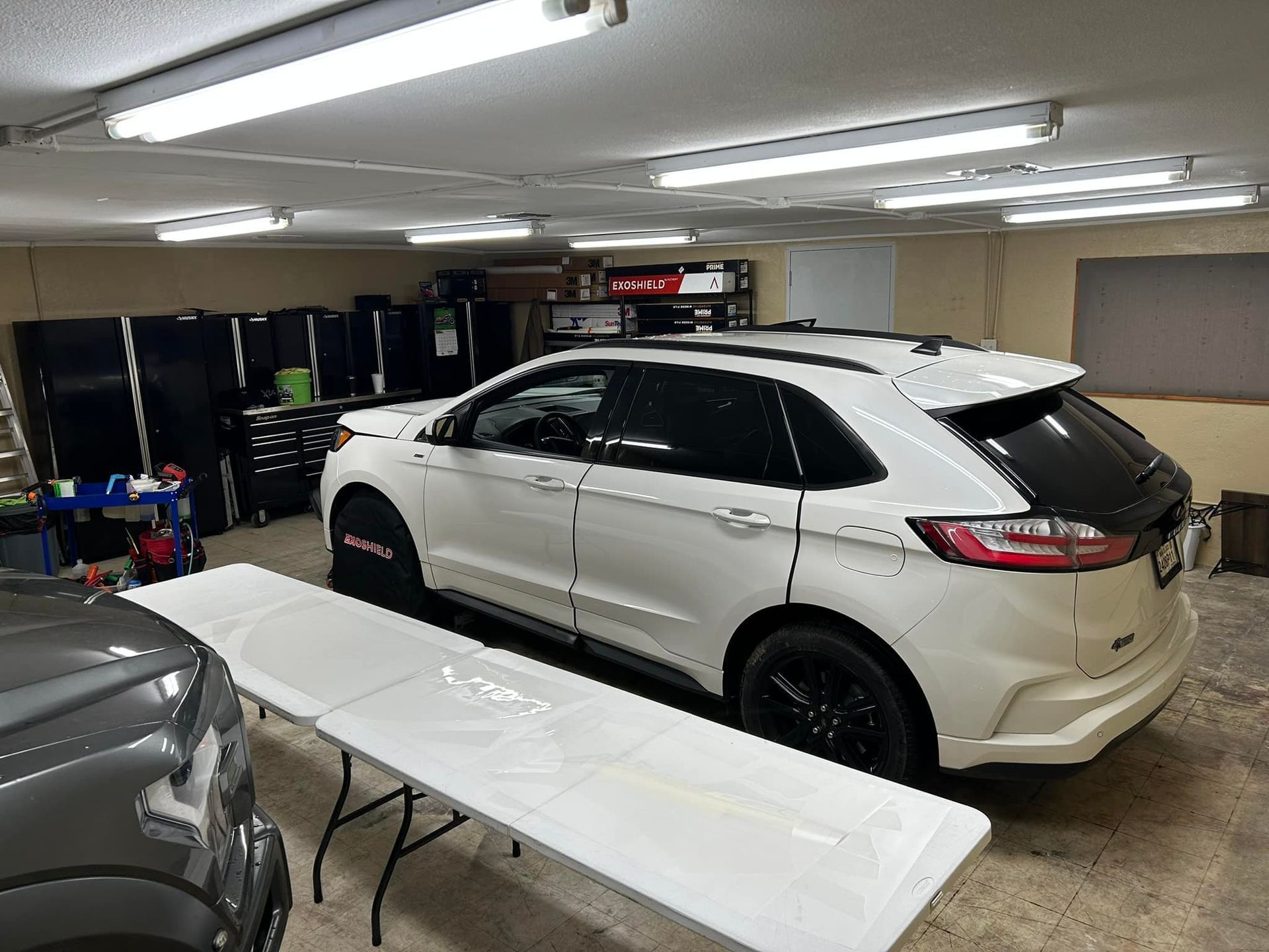 White SUV parked in a garage with a folding table in front of it and black storage cabinets.