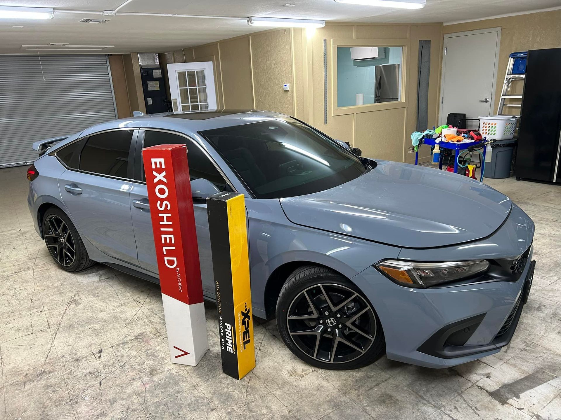 Gray Honda Civic with black rims, parked indoors, next to promotional signs.