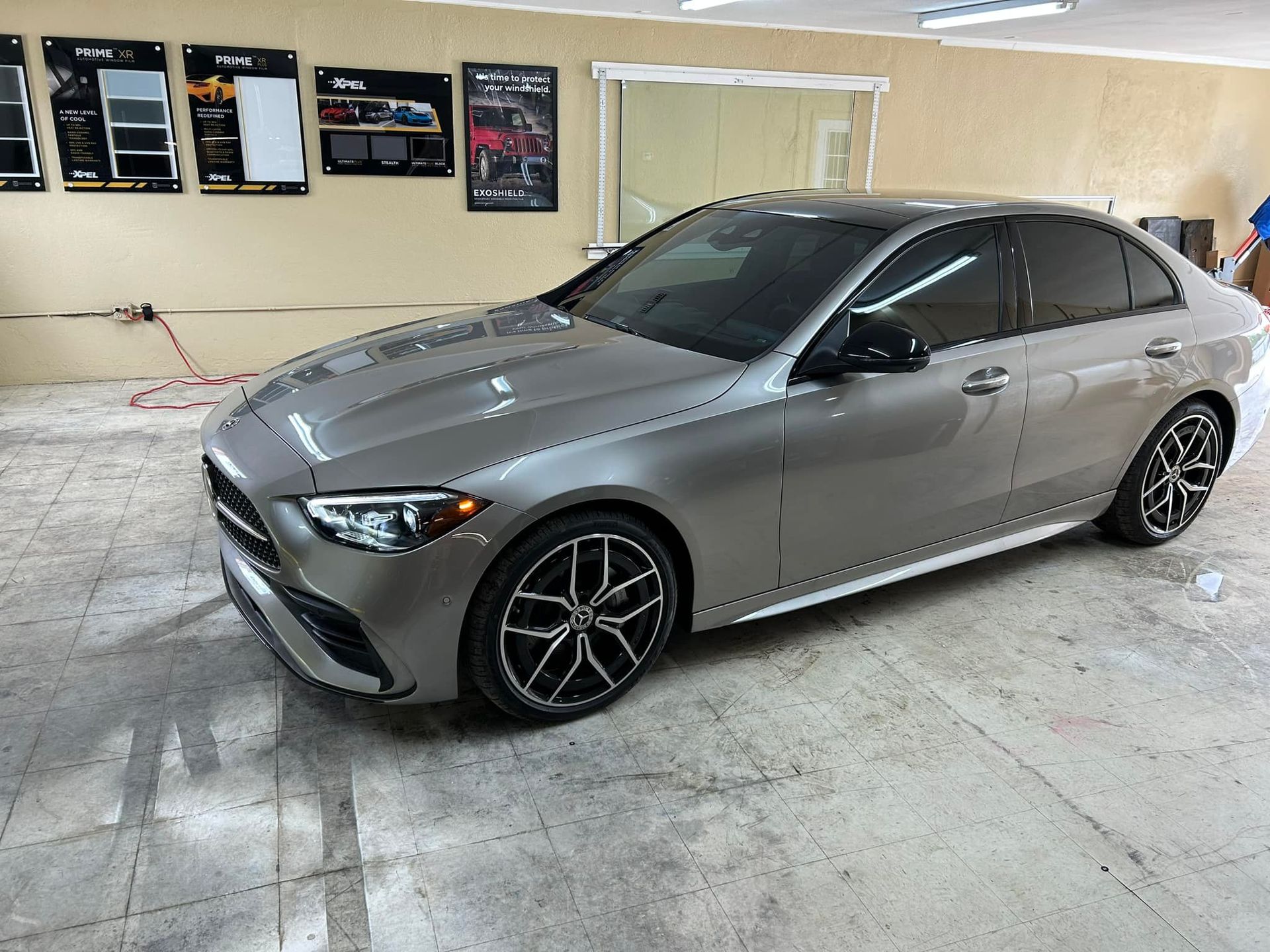 Gray Mercedes sedan parked inside a shop, windows tinted, with examples on wall in background.