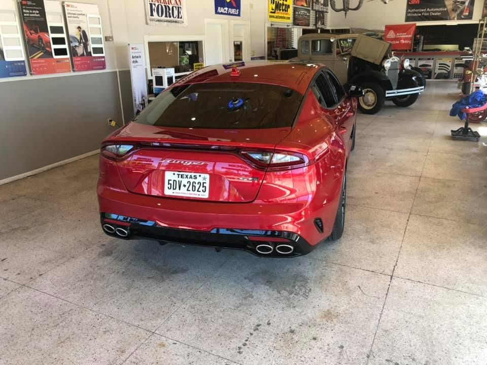 Red Kia Stinger car in a garage, Texas license plate. Other classic car and shop items are visible.