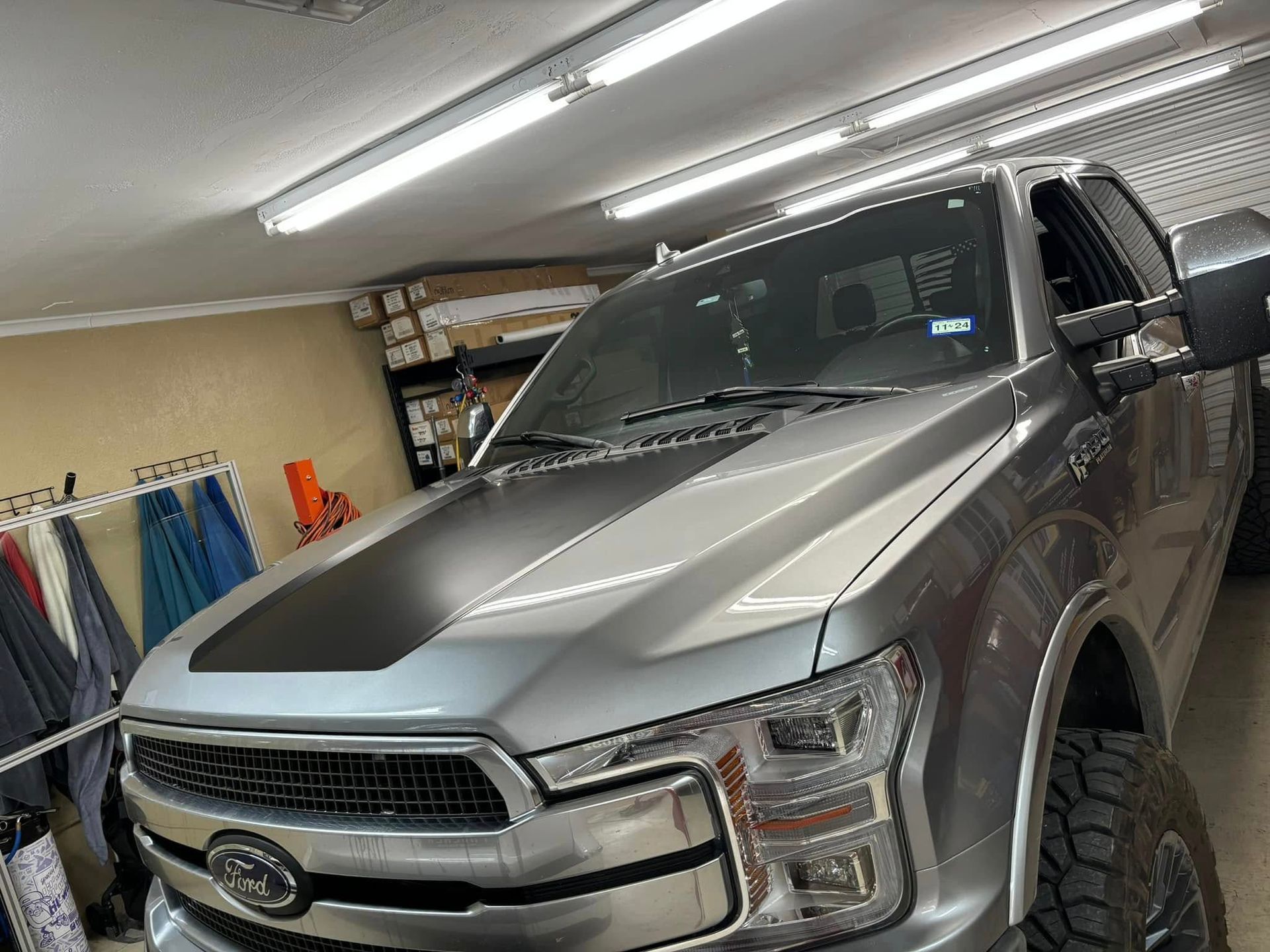 Silver Ford truck with black hood stripe parked in a garage, lit by fluorescent lights.