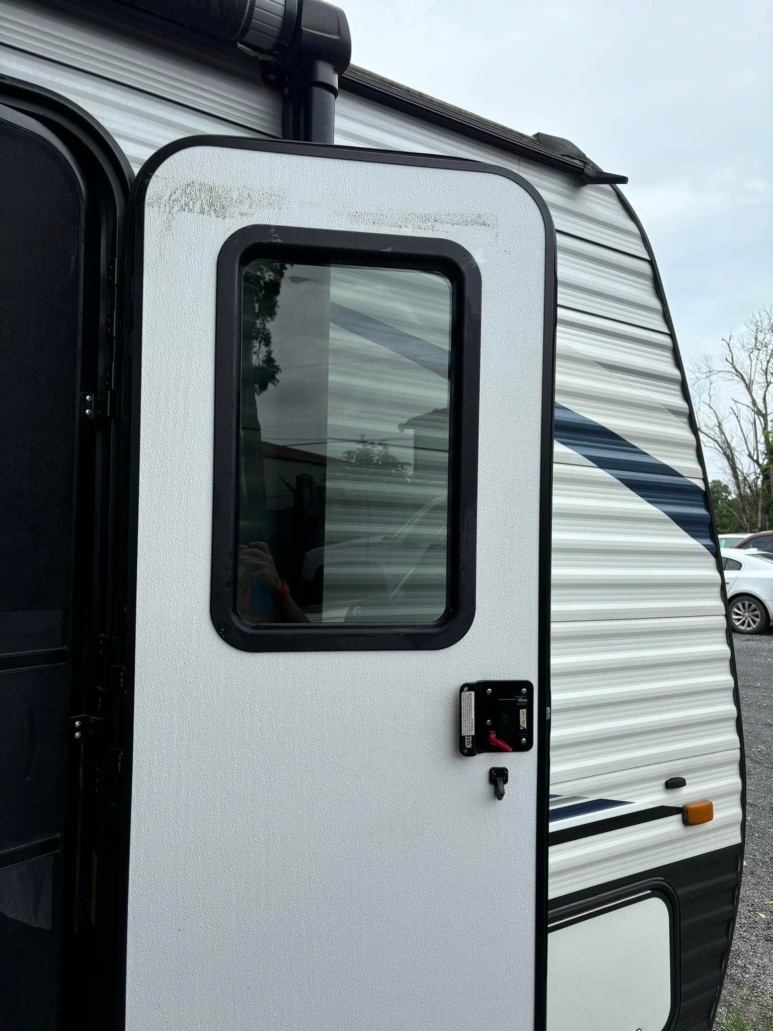 RV door with window, light gray speckled surface. Black window frame, lock. Exterior with striped siding.