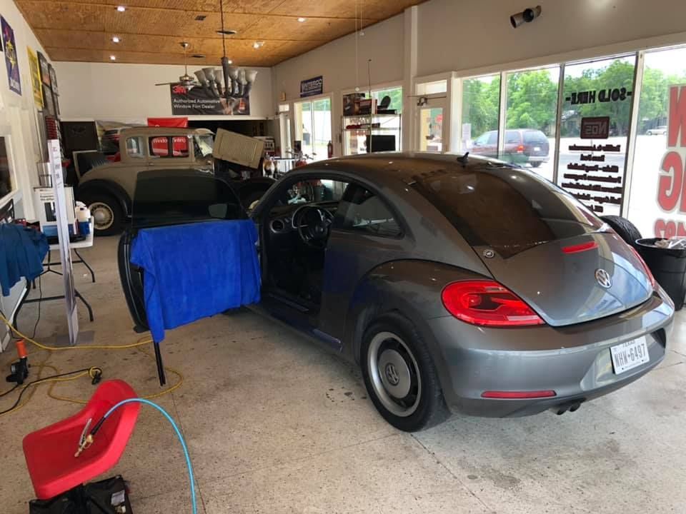 Gray VW Beetle in shop, door open, being worked on. A classic car is in the background.