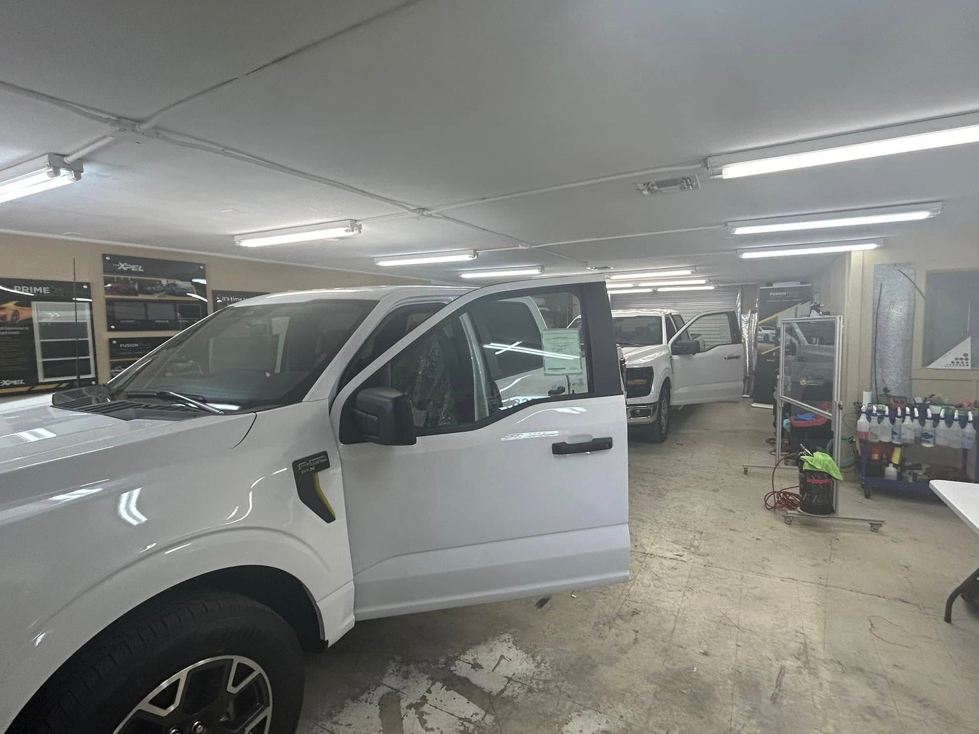 White trucks inside a garage, doors open. Lighting, tools, and shelves are visible.