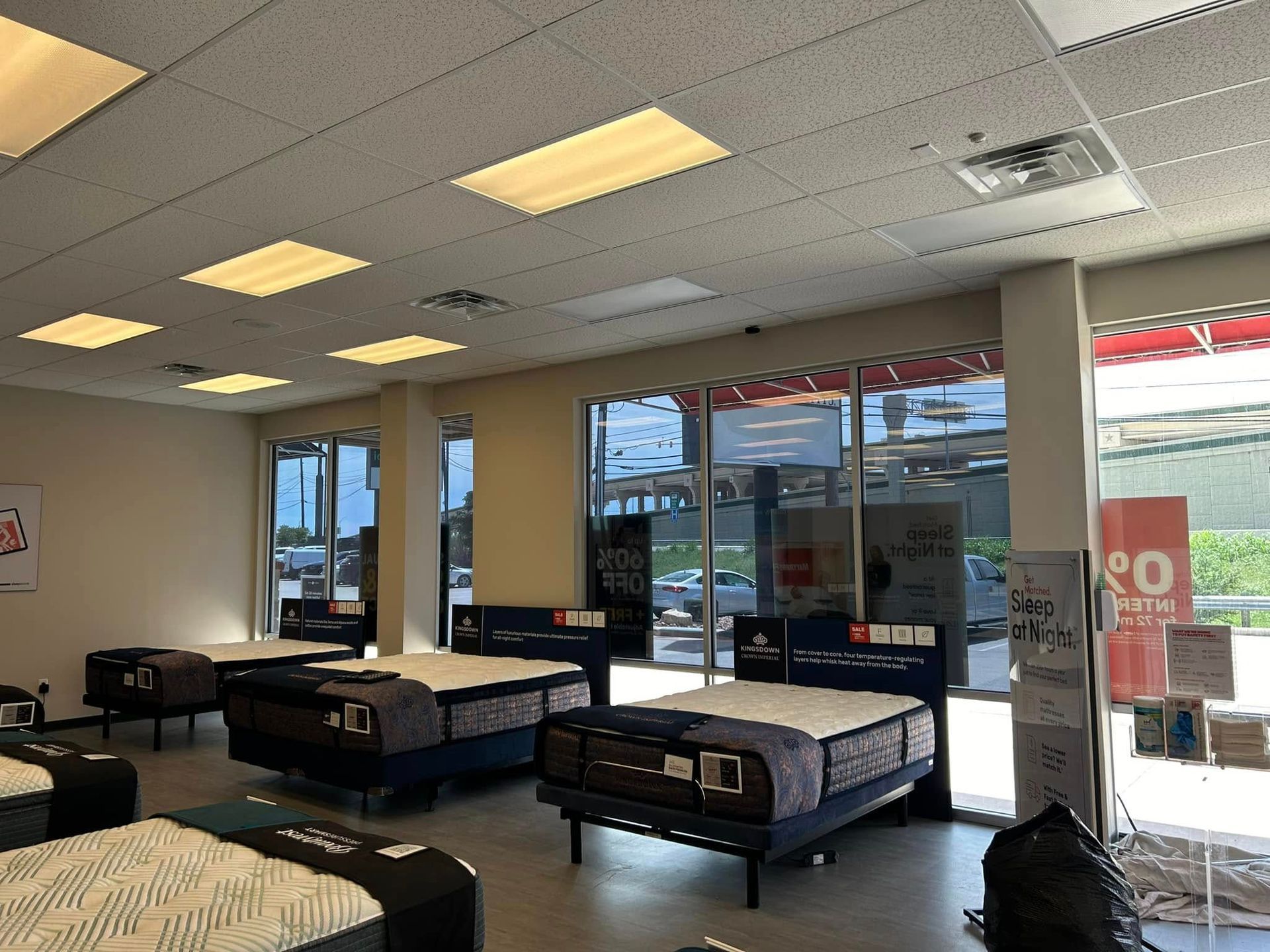 Mattress store interior: Multiple mattresses on display, lit by overhead lights, with large windows visible.