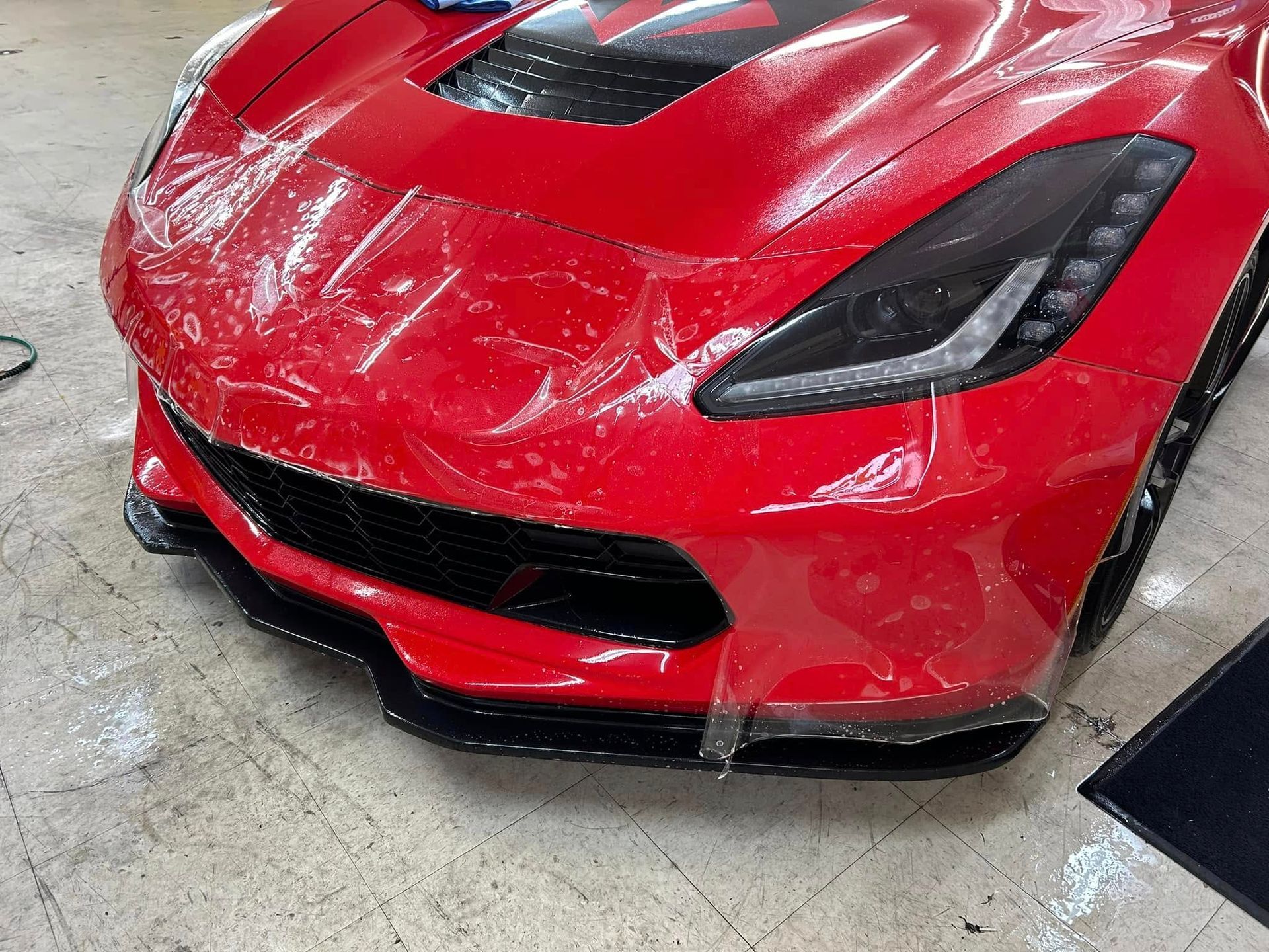 Red Corvette with protective film being applied to its front bumper in a garage.