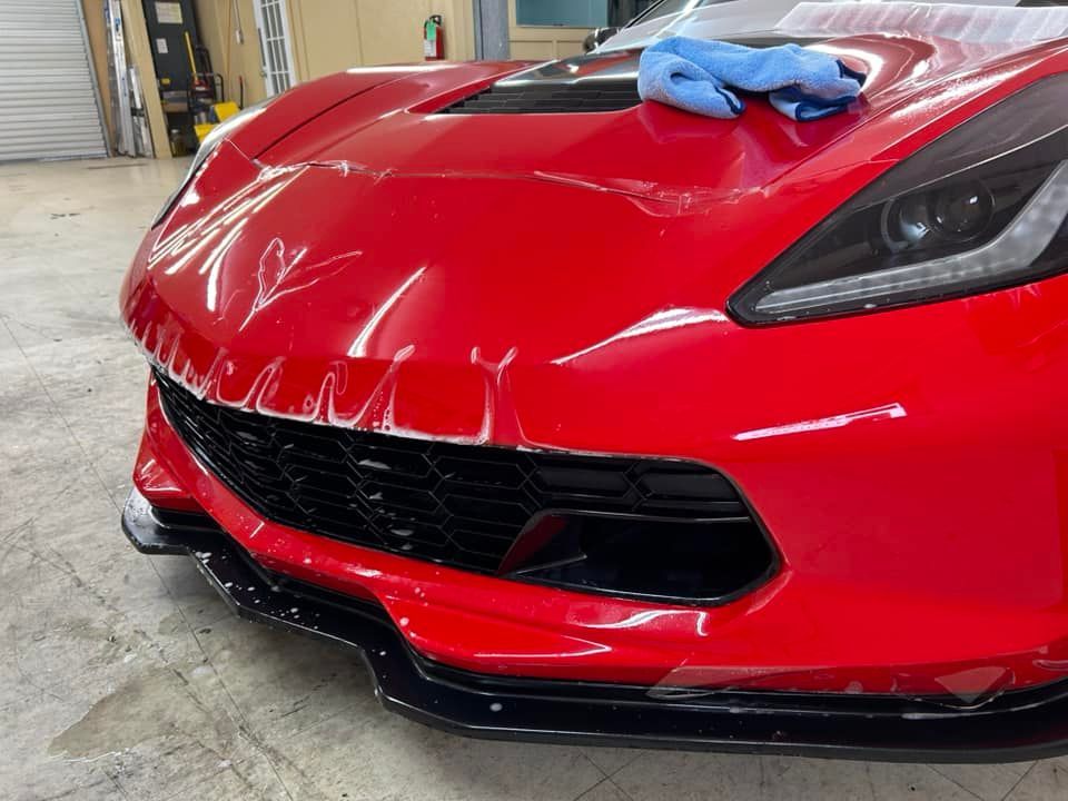 Red Corvette with protective film being applied to the front bumper in a garage.
