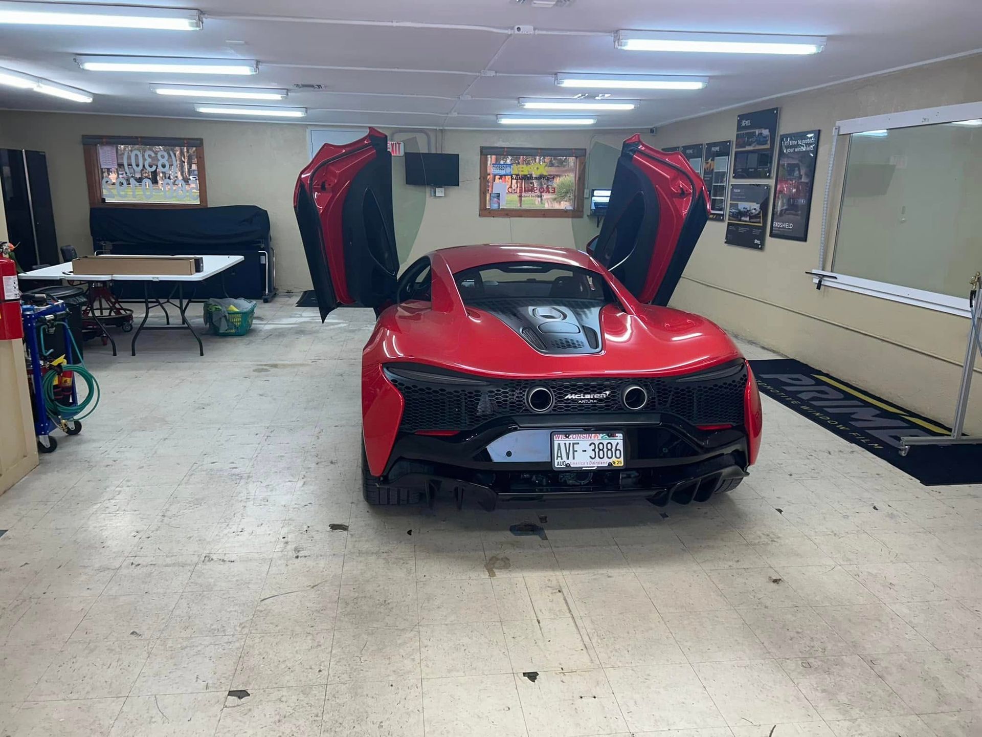 Red sports car with doors open in a garage, under fluorescent lights.