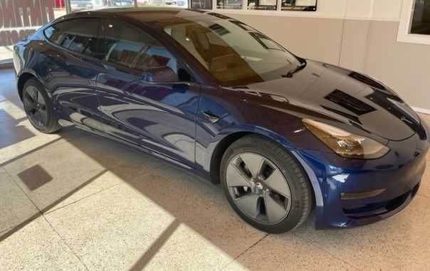 Blue Tesla Model 3 parked inside a building with light brown flooring, angled towards the camera.