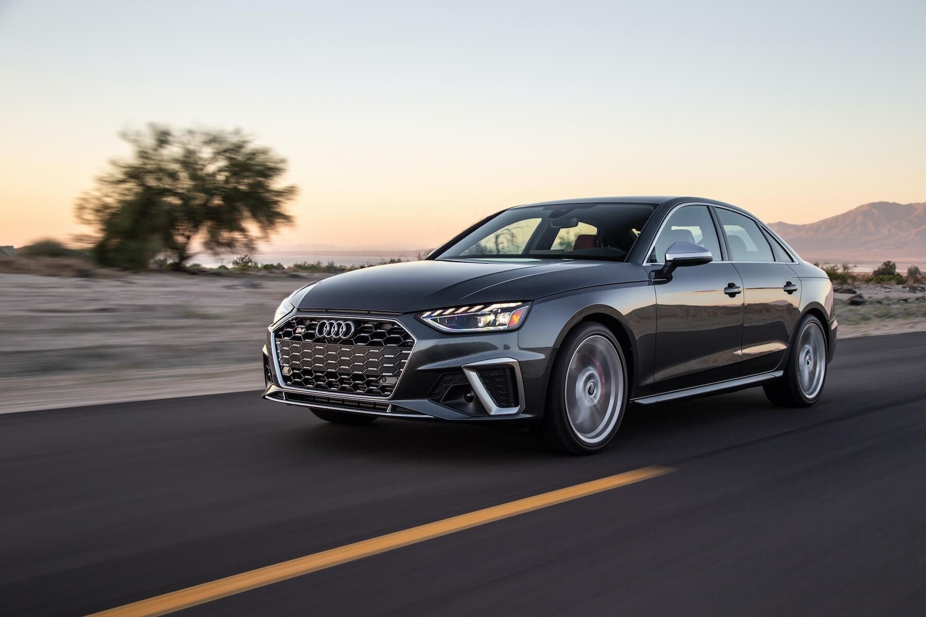 Gray Audi S4 sedan driving on a road with desert scenery and sunset.