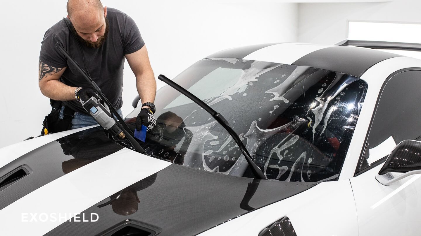 Man applying protective film to a white car windshield. Inside a shop, applying fluid and smoothing.