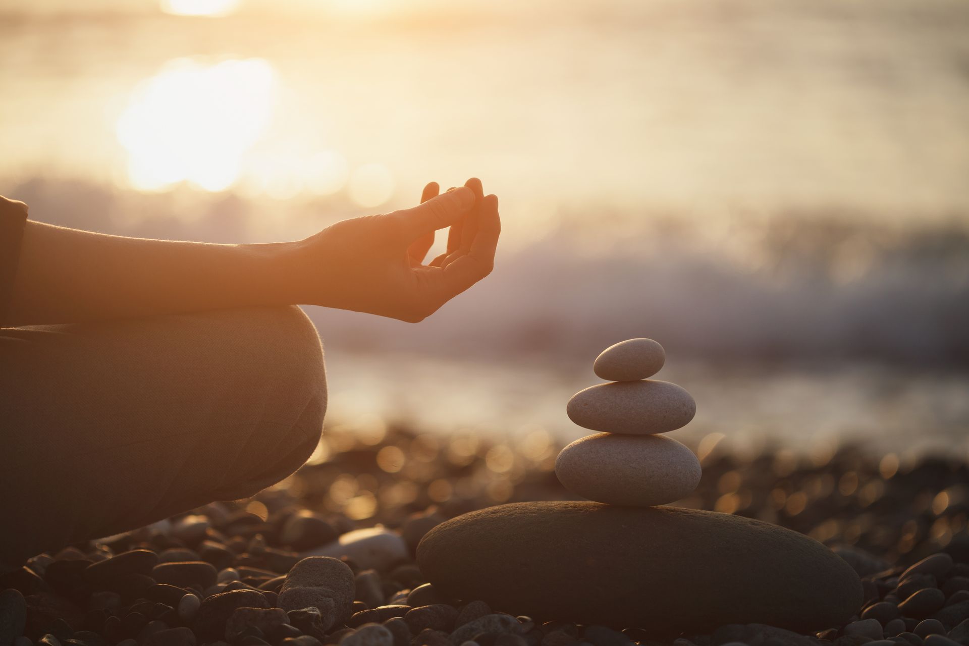 A person is meditating on the beach next to a stack of rocks.
