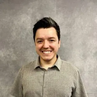 Man with dark hair and a friendly smile wearing a gray button-down shirt against a gray background.