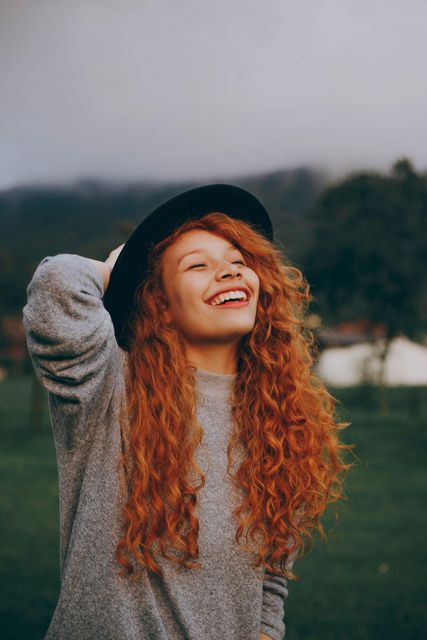 A woman with long red hair is wearing a hat and smiling.