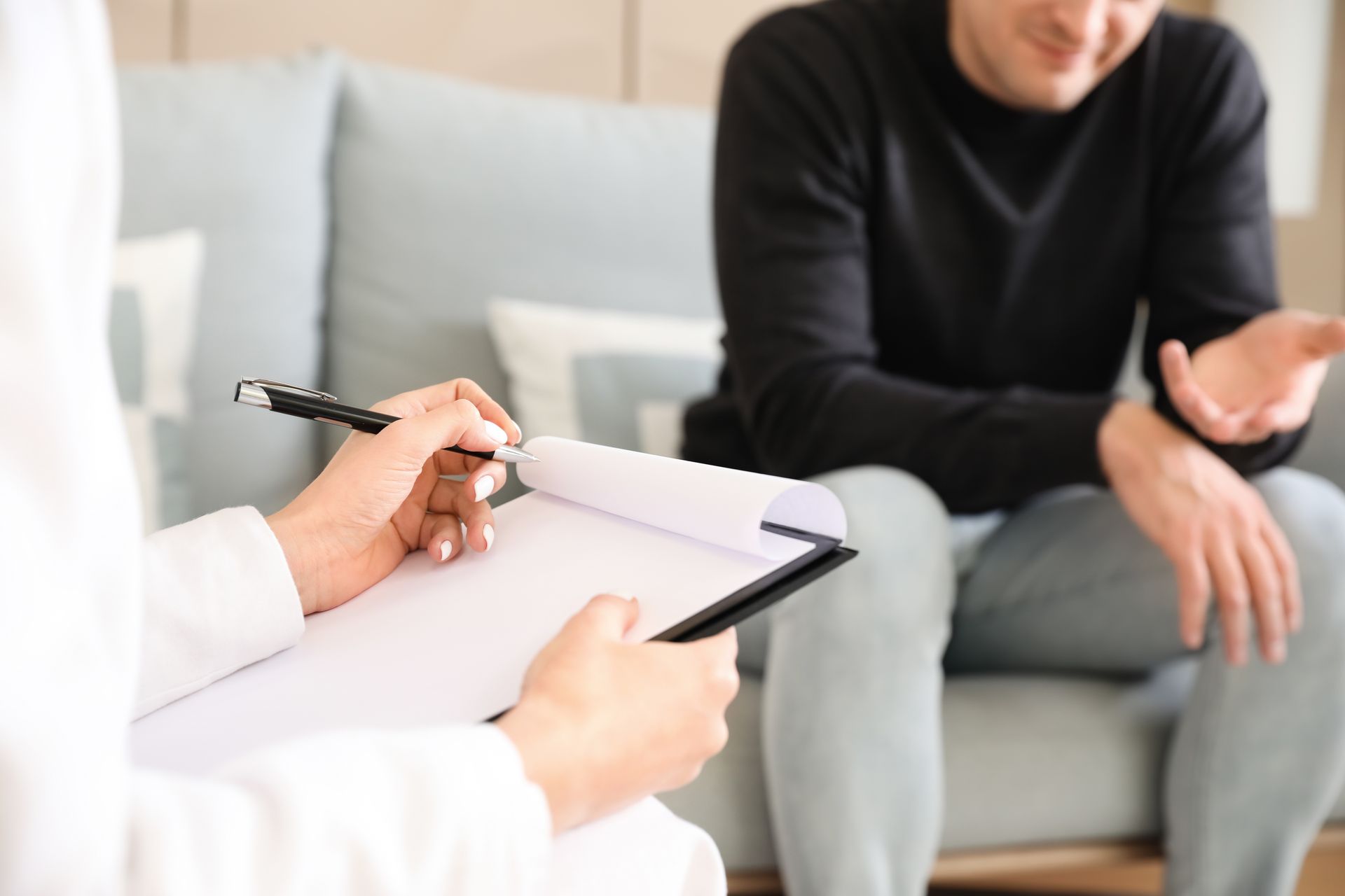 A therapist in a white coat takes notes on a clipboard while a person sits on a sofa, gesturing during a consultation.