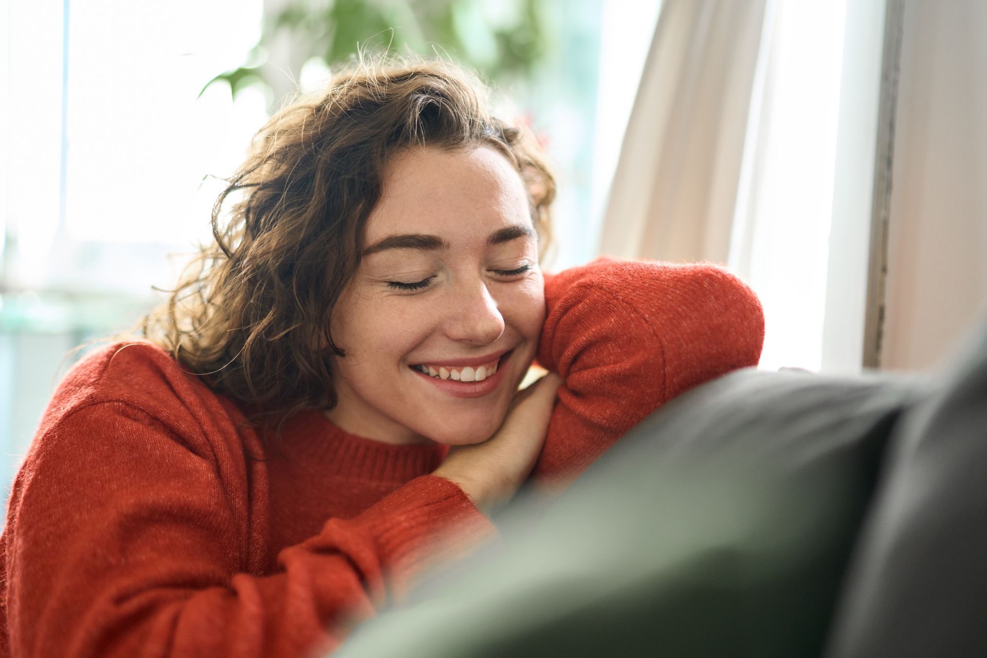 Woman with curly hair smiles, leaning on a sofa, wearing a red sweater indoors.