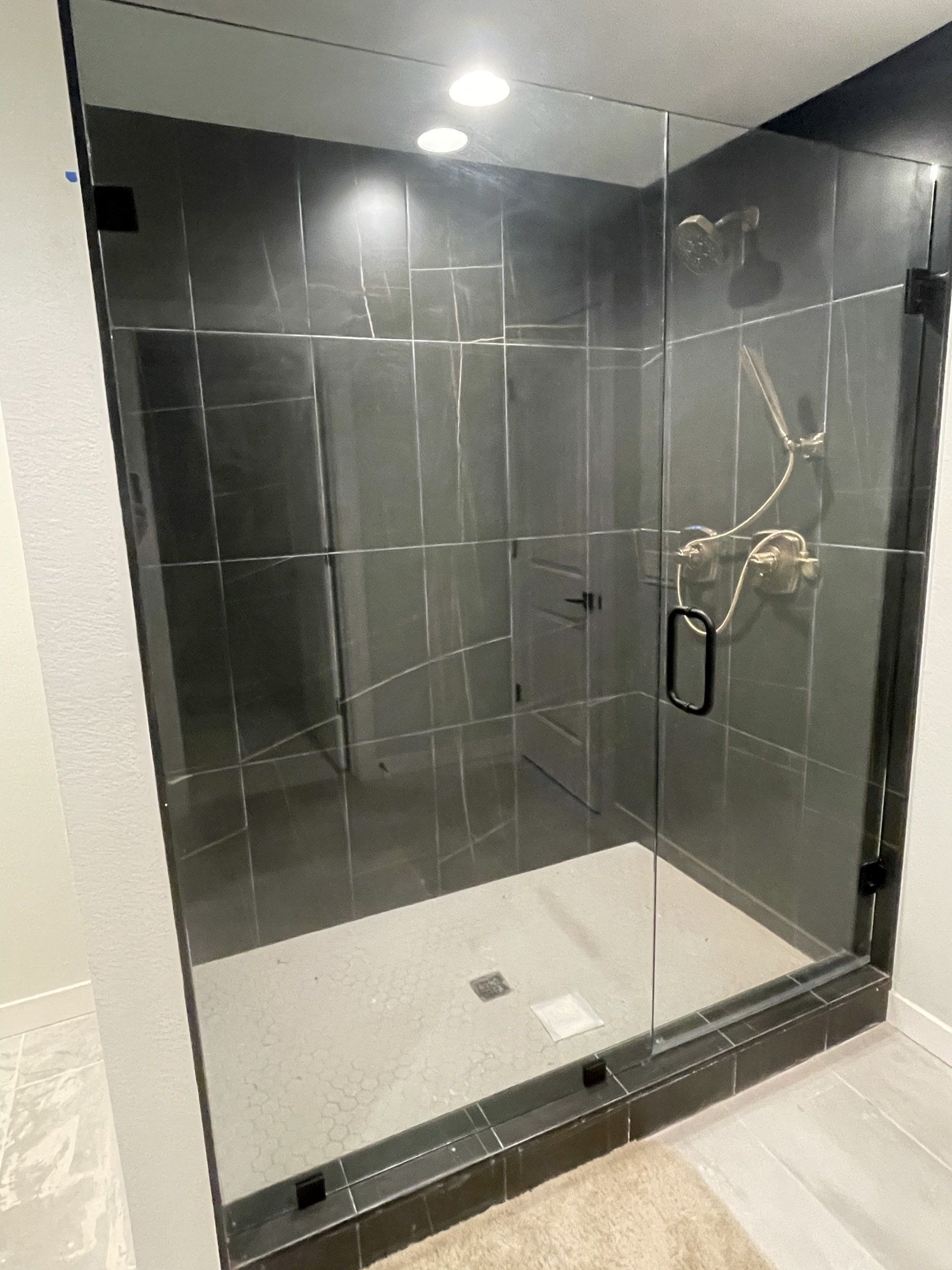 Glass-enclosed shower with dark gray tile walls and white floor. Silver shower fixtures and black door handle.