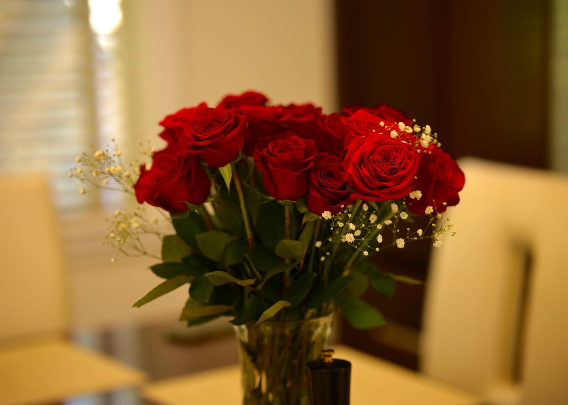 Bouquet of red roses in a glass vase, set on a table with chairs, inside a room.