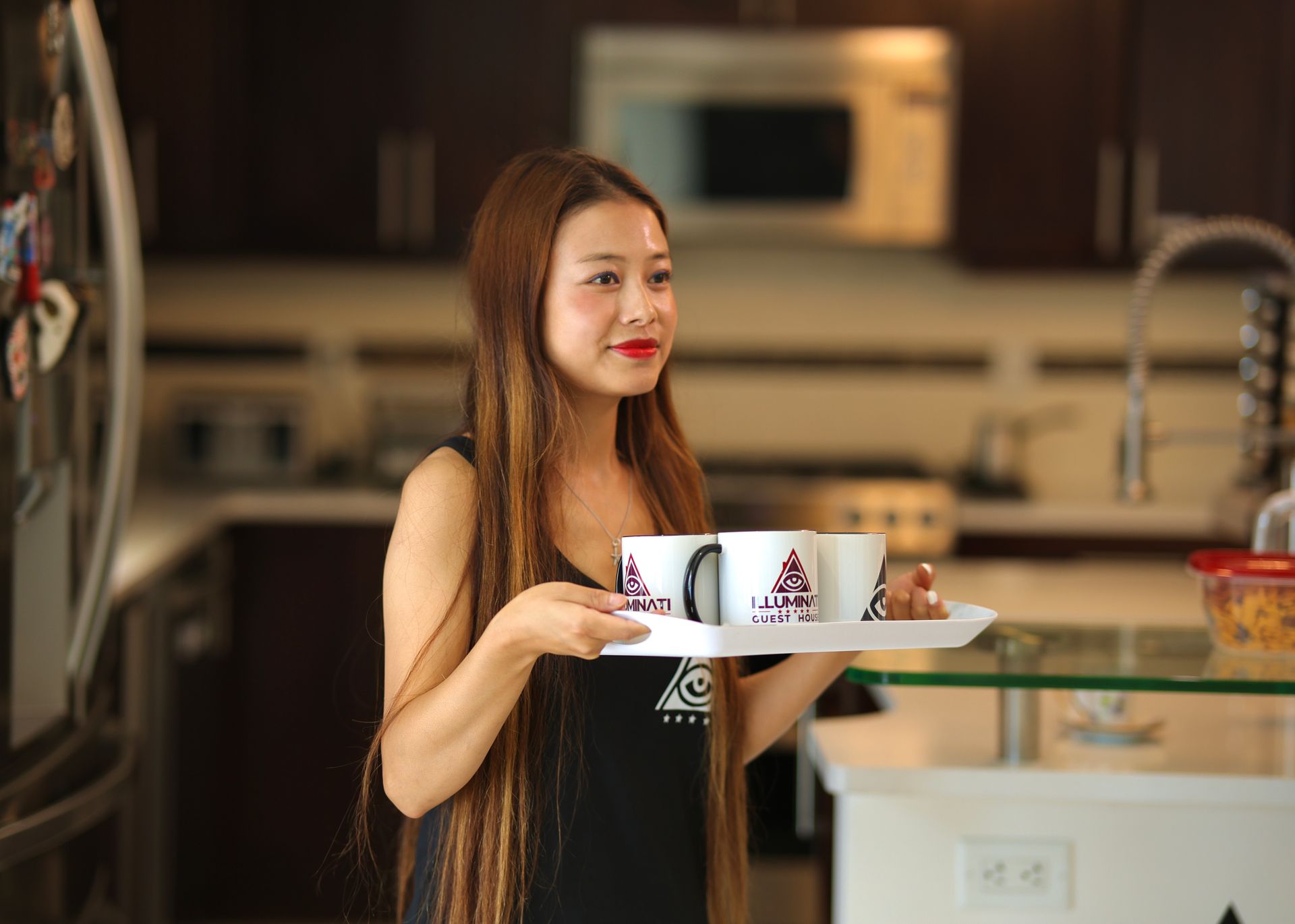 Woman in kitchen holding tray with two mugs.