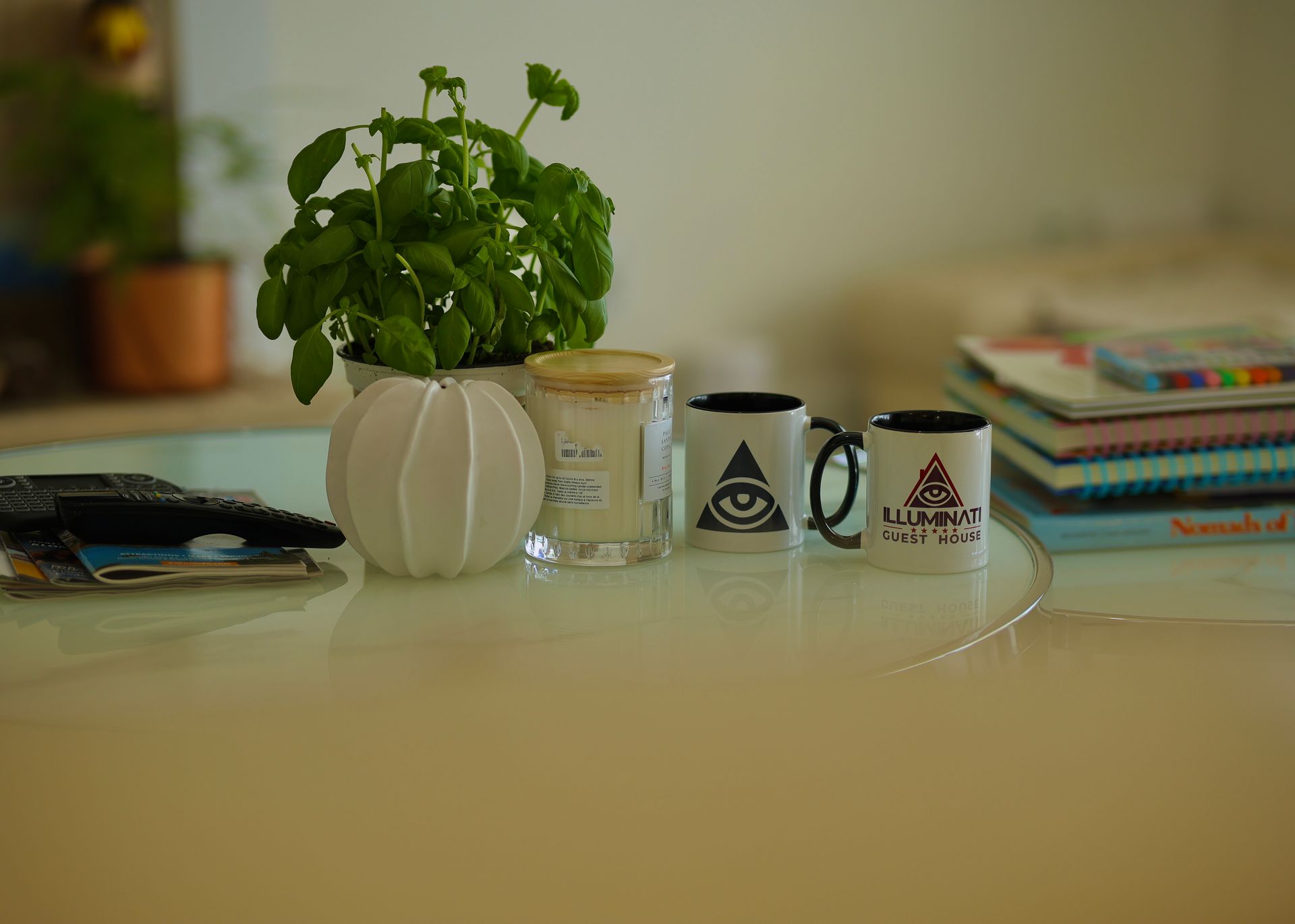 A tabletop scene: basil plant, candle, two mugs with eye symbols, books.