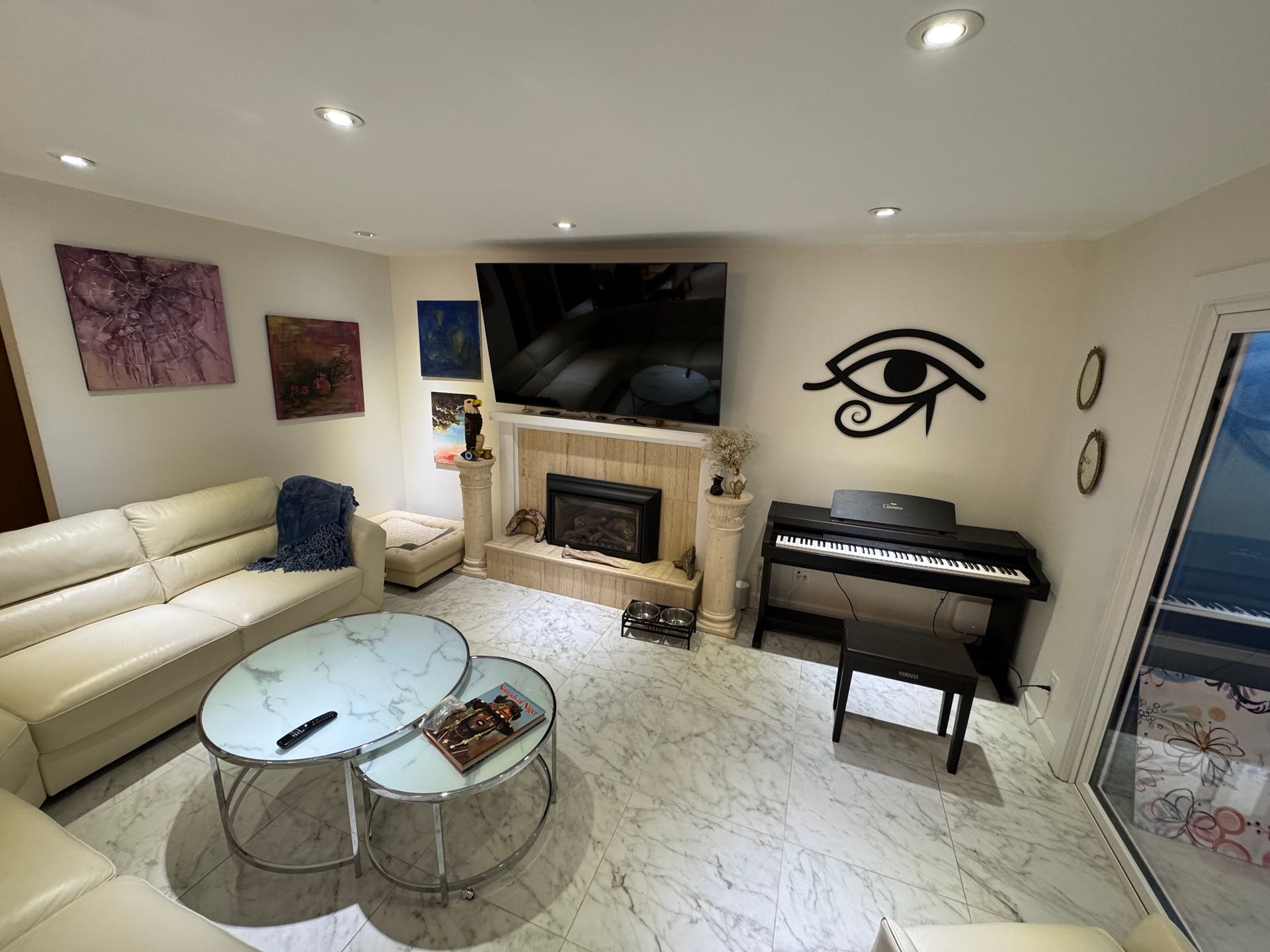 Living room with cream sofa, marble floors, fireplace, piano, and art. Wall features a large black Eye of Horus.