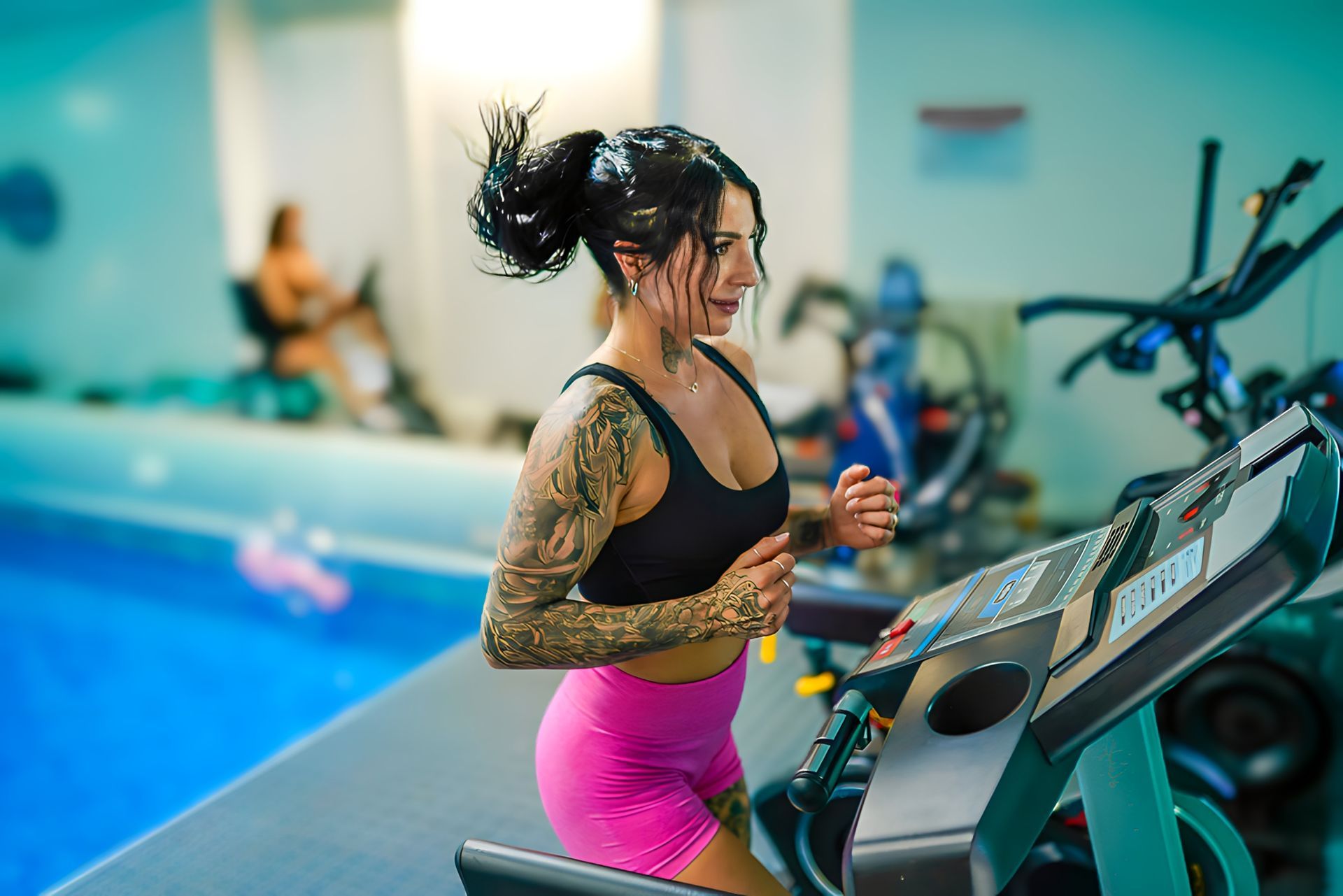 Woman with tattoos runs on a treadmill by a pool, wearing pink shorts and black top.