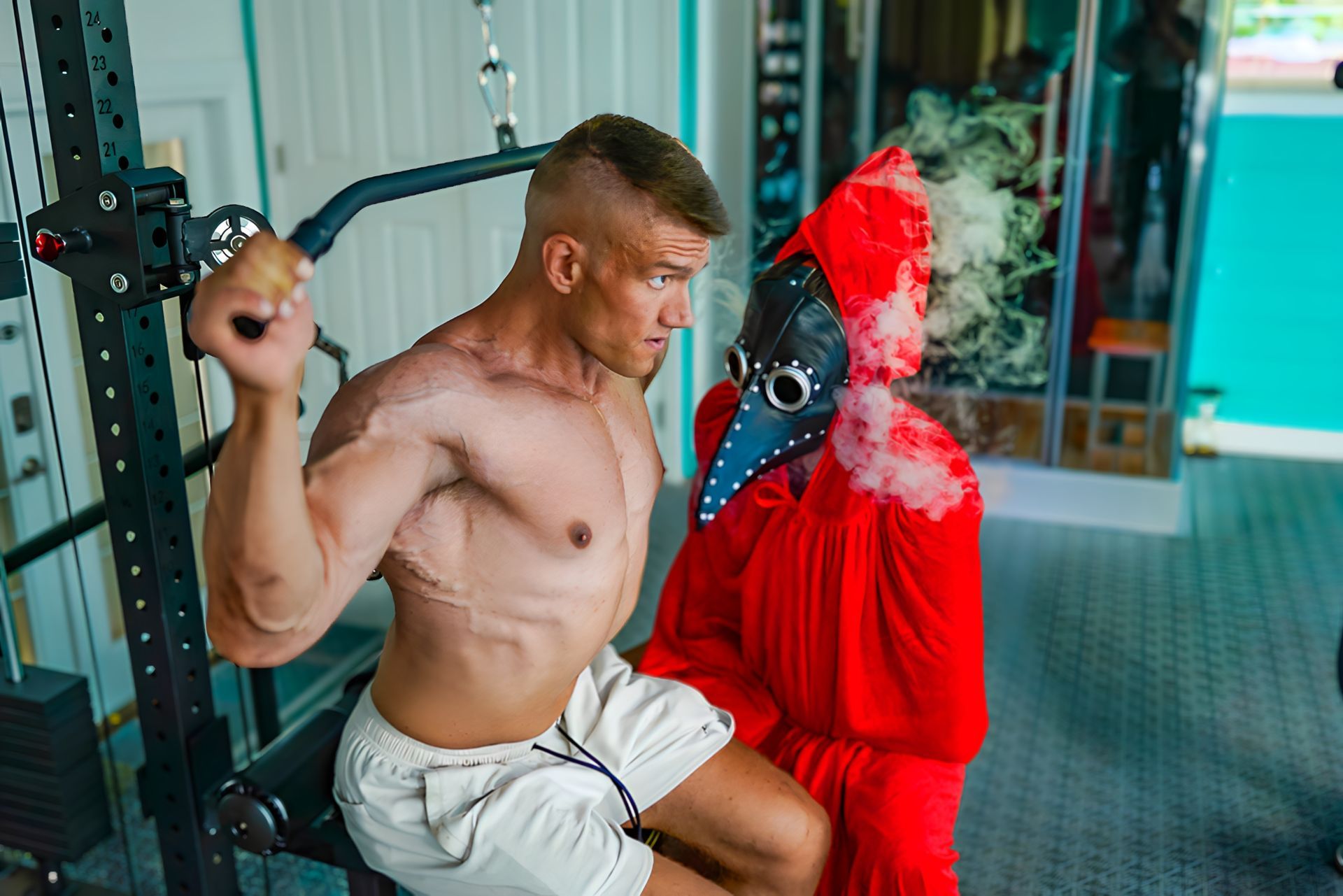 A man in a plague doctor mask is sitting next to a shirtless man in a gym.