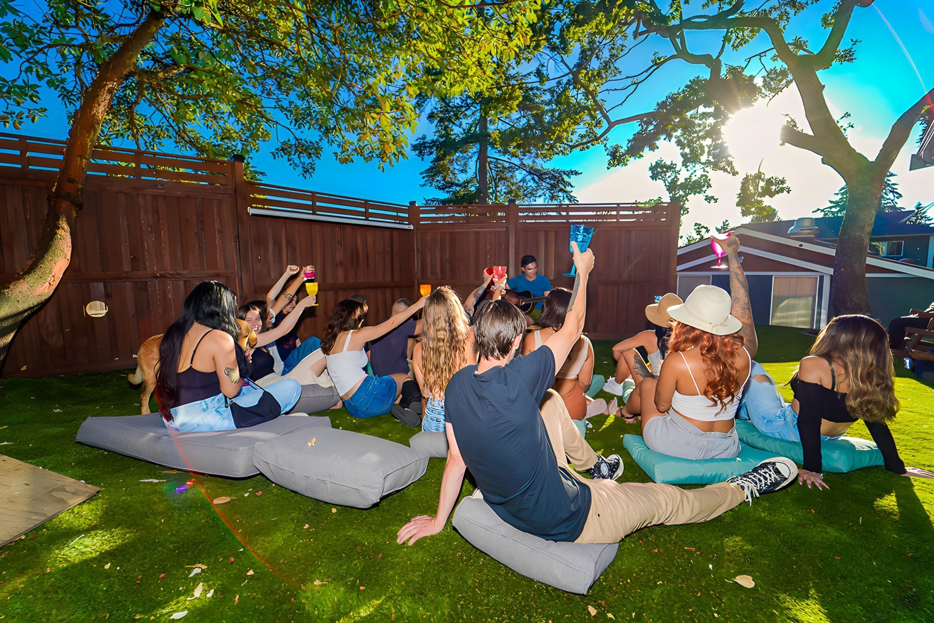 A group of people are sitting on pillows on the grass in a backyard.