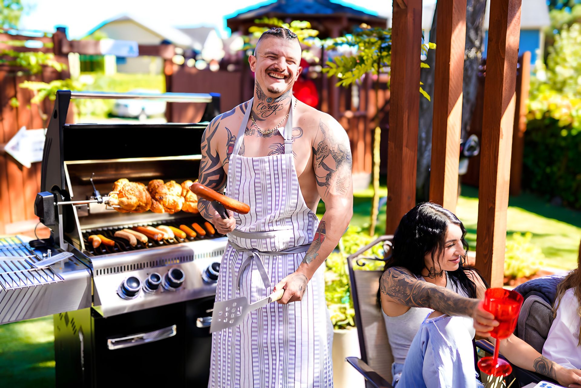 Man grilling in backyard, wearing apron, smiling, with tattoos. Others seated nearby. Sunny day.