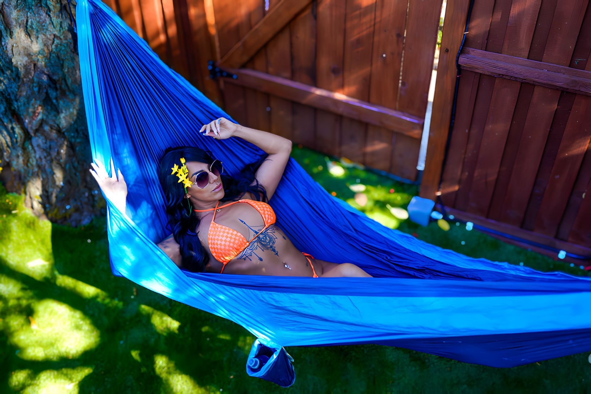 A woman in a bikini is laying in a blue hammock in front of a wooden fence.