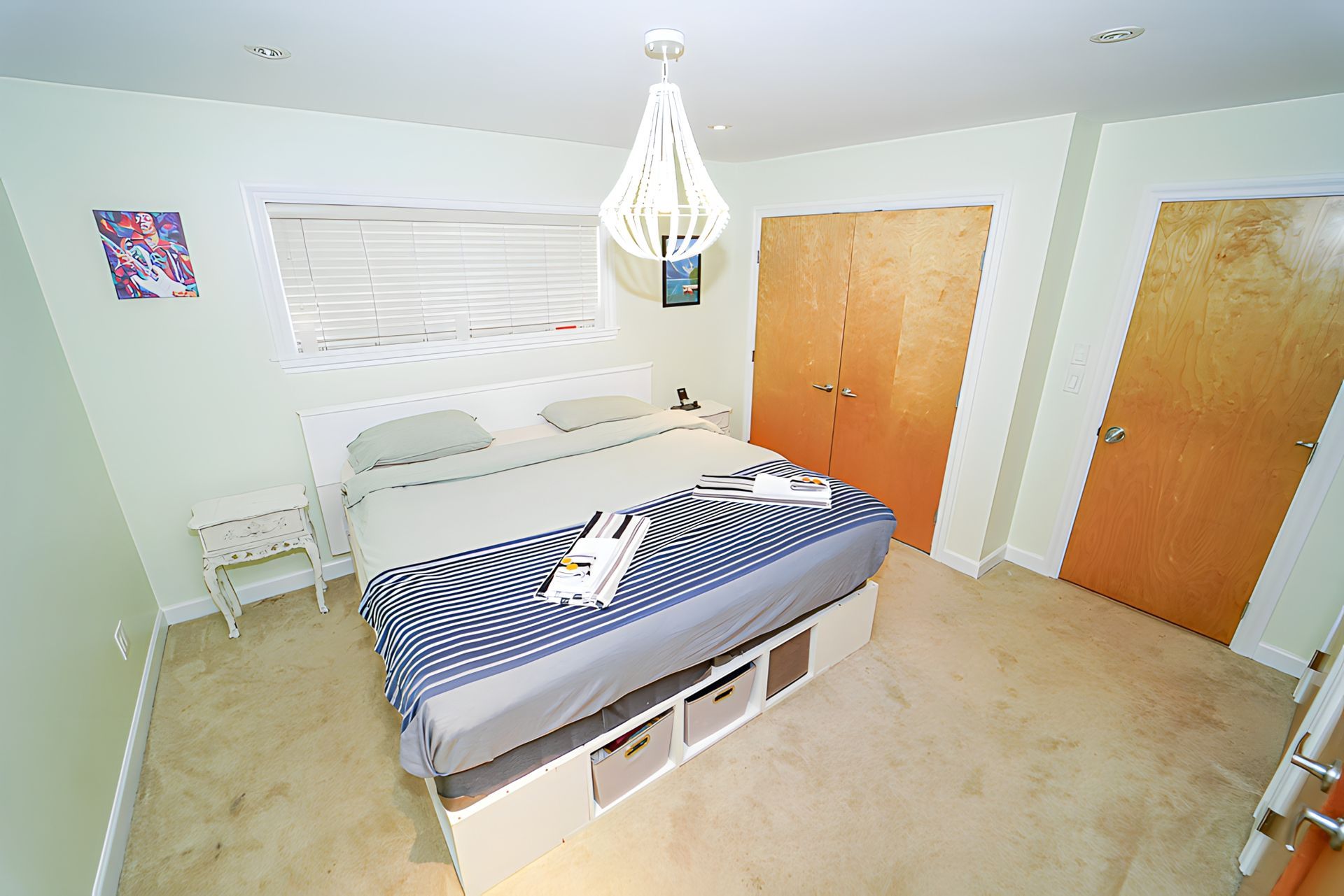 Bedroom with white bed, beige carpet, two wooden doors, a window, and a white chandelier.