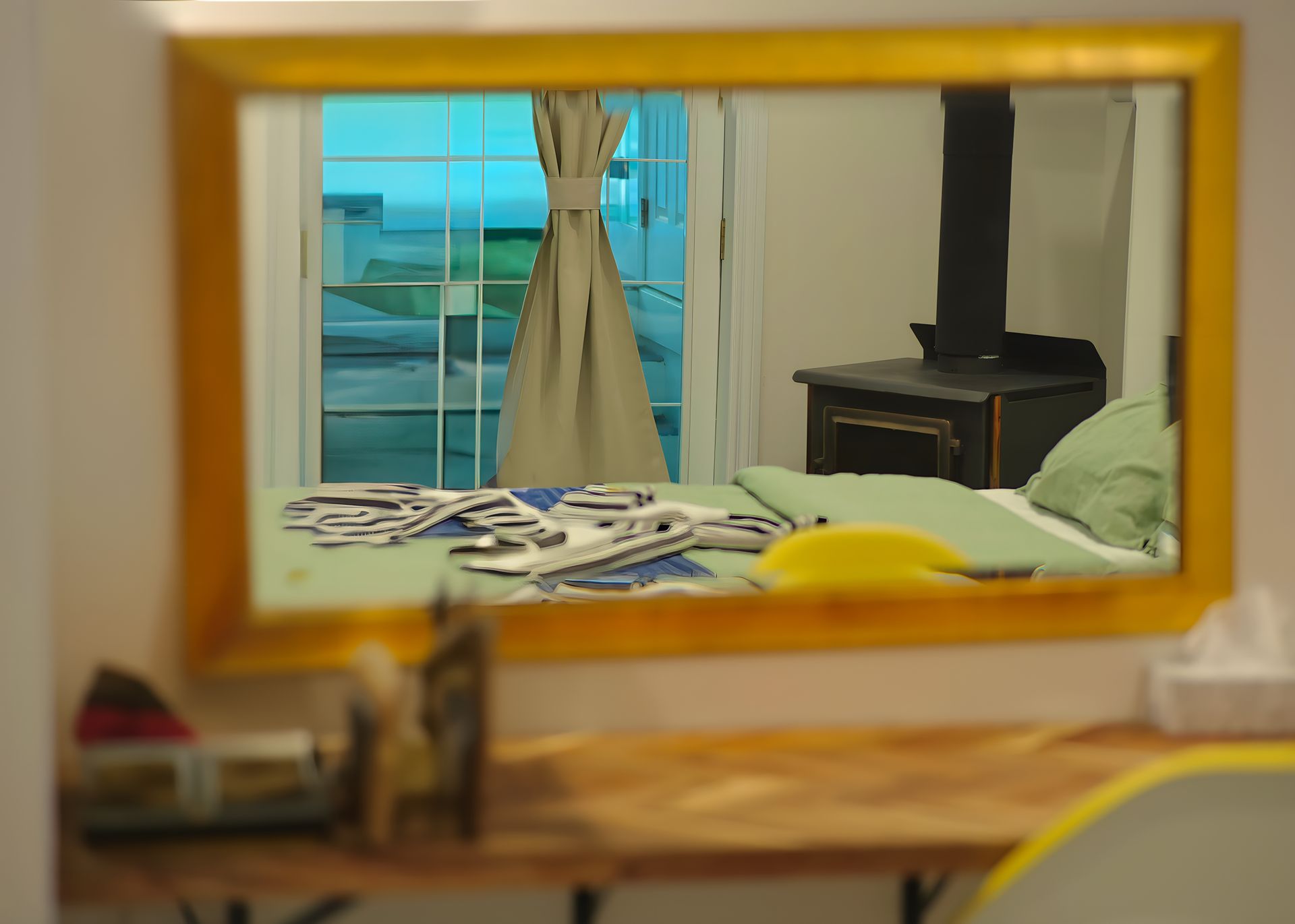 Mirror reflecting a bedroom with a window, bed, and dresser. The mirror has a gold frame.