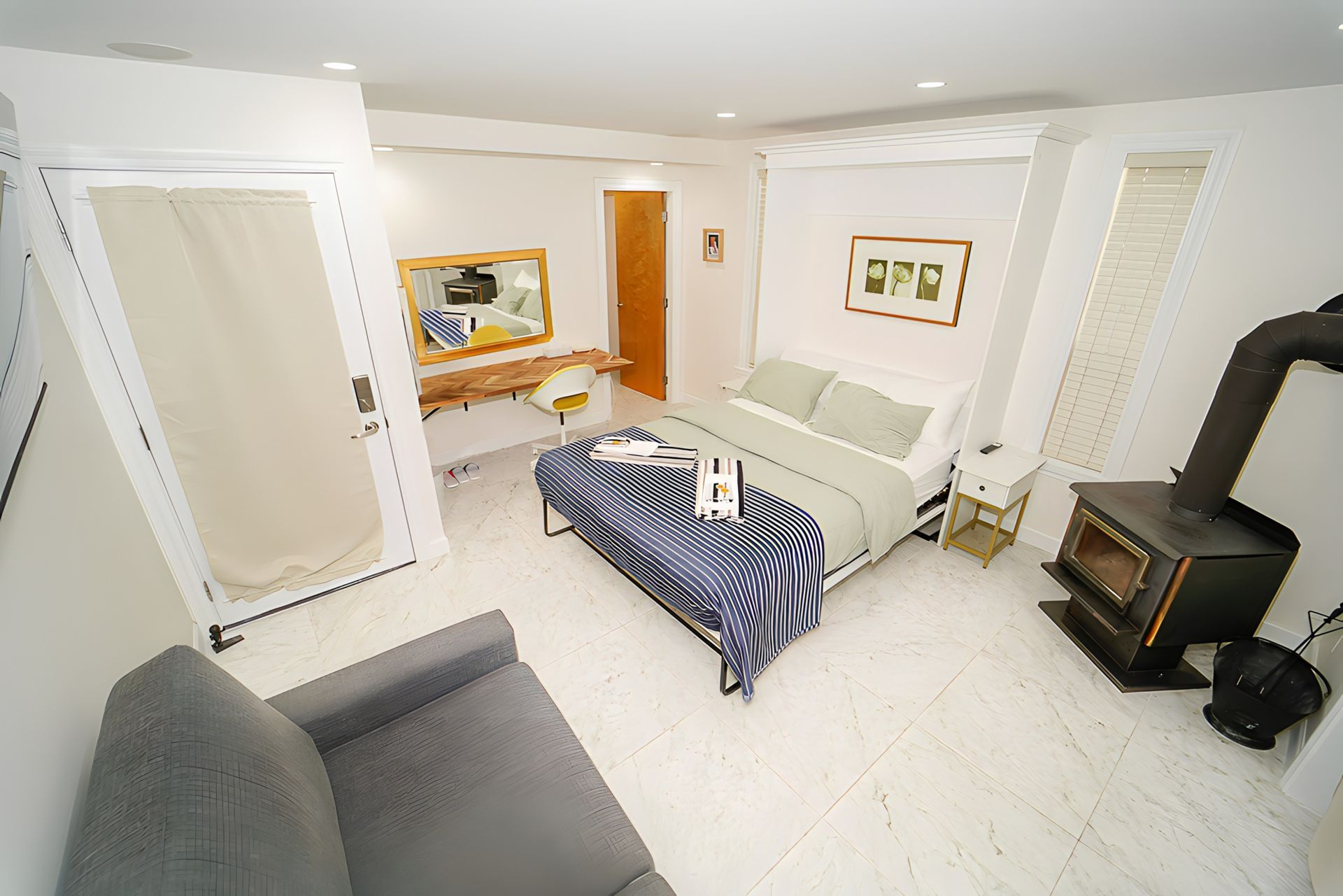 Bright, white bedroom with bed, couch, desk, wood stove. Window with white blinds.