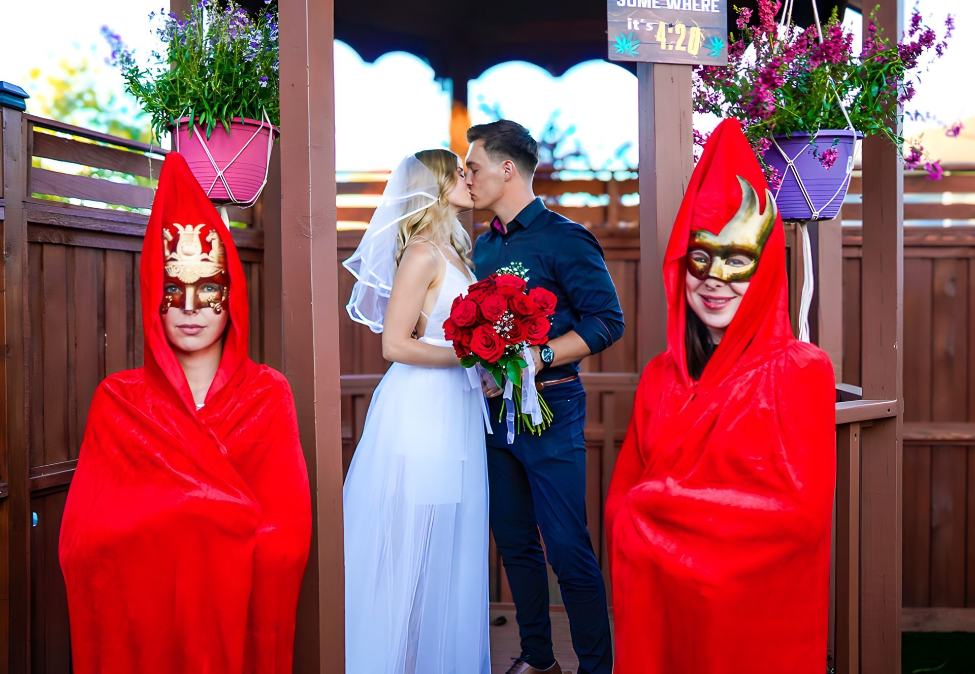 A bride and groom are kissing in front of a gazebo while two people in red cloaks are standing behind them.