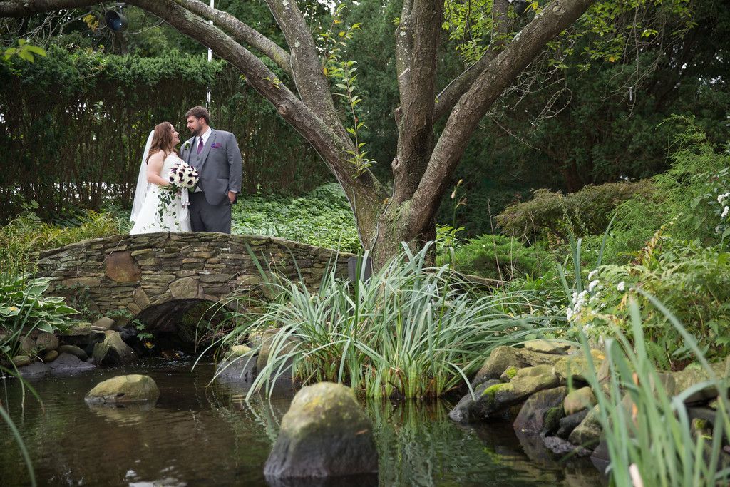 A bride and groom are standing on a stone bridge over a pond.