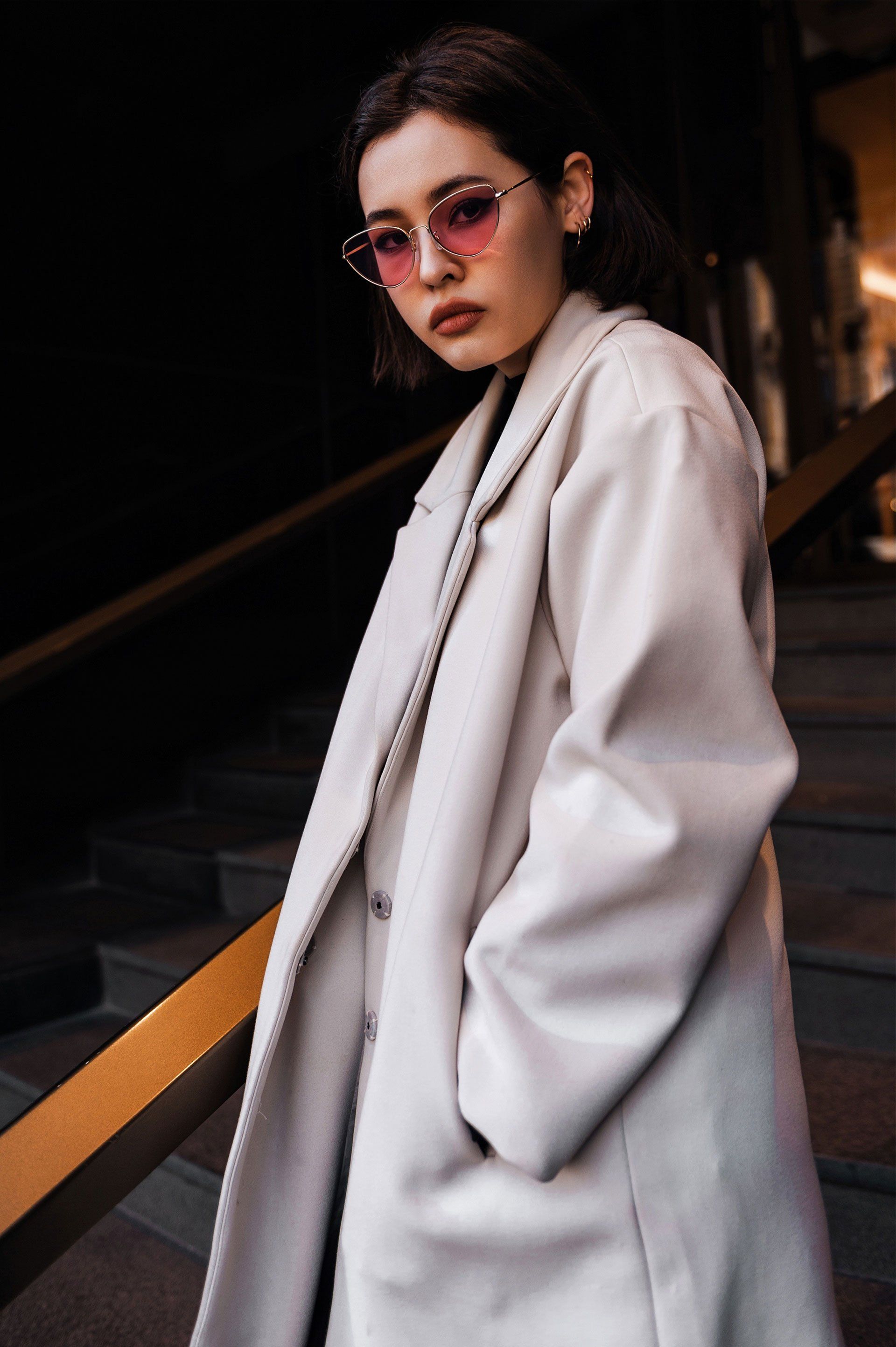 Una mujer con un abrigo claro y gafas de sol rosas está parada en las escaleras, con las manos en los bolsillos, mirando a la cámara.