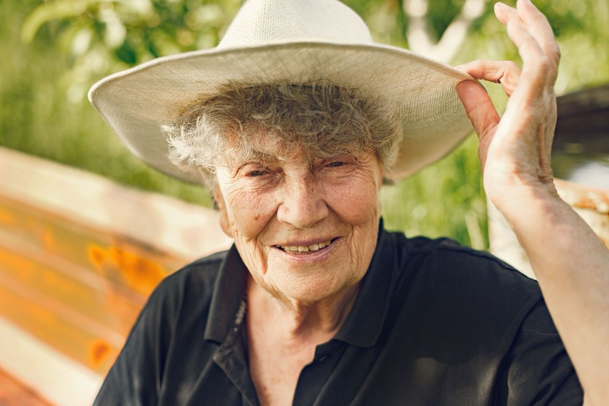 Smiling person in a straw hat and black shirt outdoors, holding the hat brim.