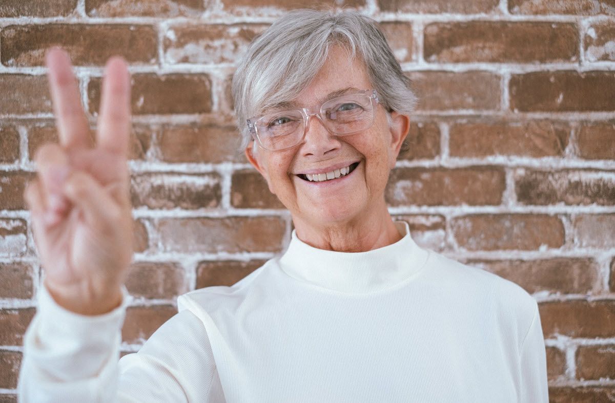 An elderly woman is giving a peace sign in front of a brick wall.