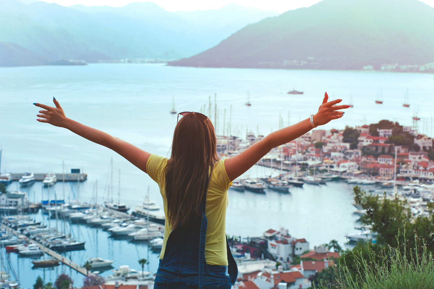 A young traveller spreads her arms wide on a hilltop, looking at a fishing town below.