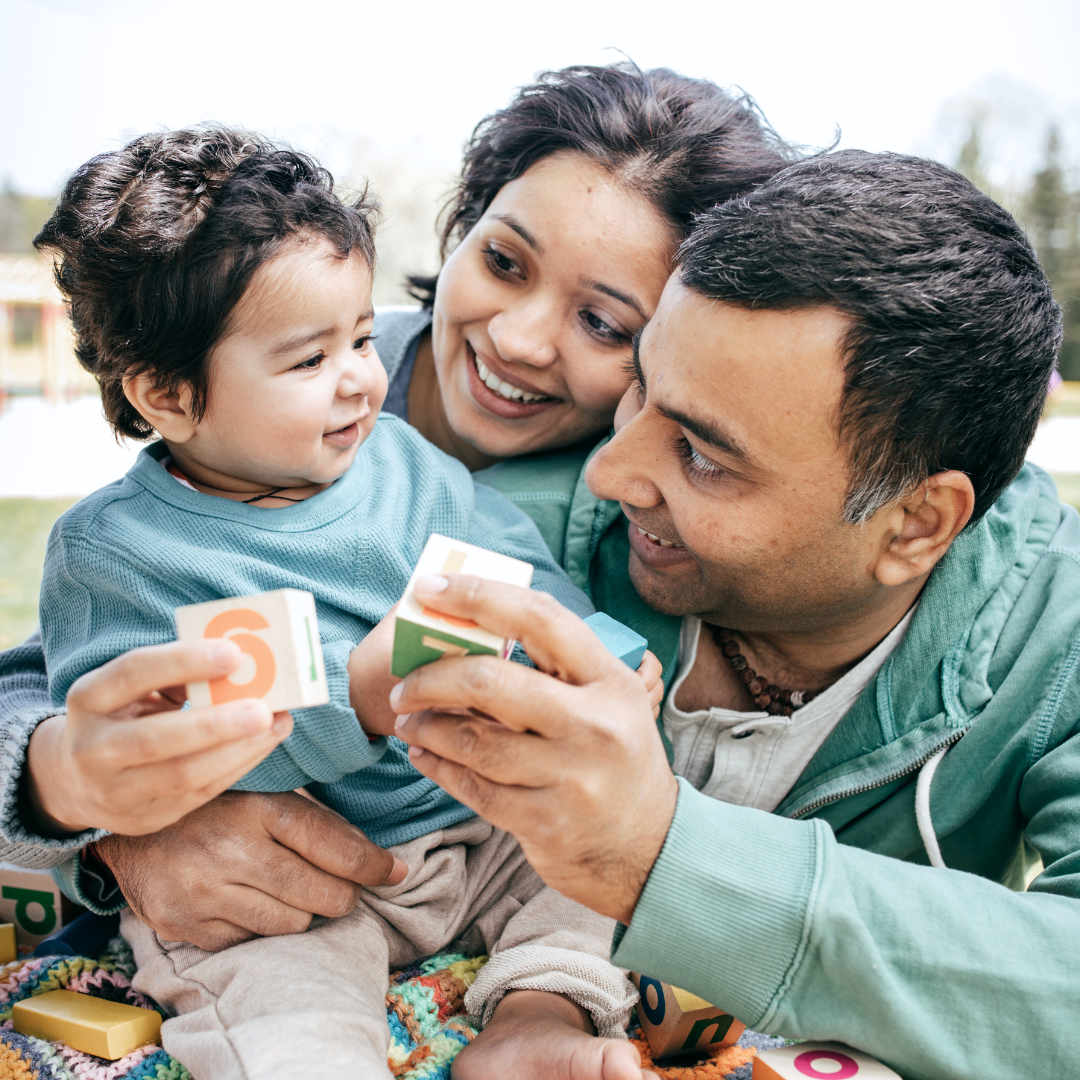 A man and woman are playing with a baby who is holding a block with the letter s on it.
