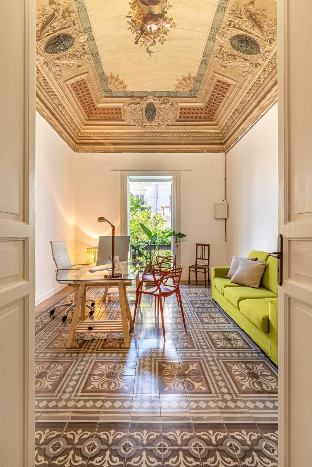 Stanza con soffitto decorato, pavimento in piastrelle decorate, scrivania, divano verde e porta aperta.