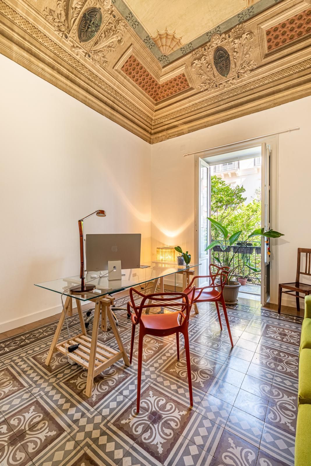 Uno studio in casa con scrivania, sedie rosse e un balcone con verde. Soffitto decorato e pavimento piastrellato.